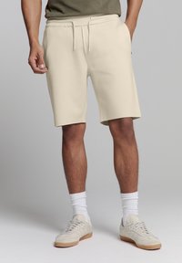Beige sportshorts gemaakt van zachte stof, met een elastische tailleband en koord, gedragen met witte crew-sokken en lichte sneakers.