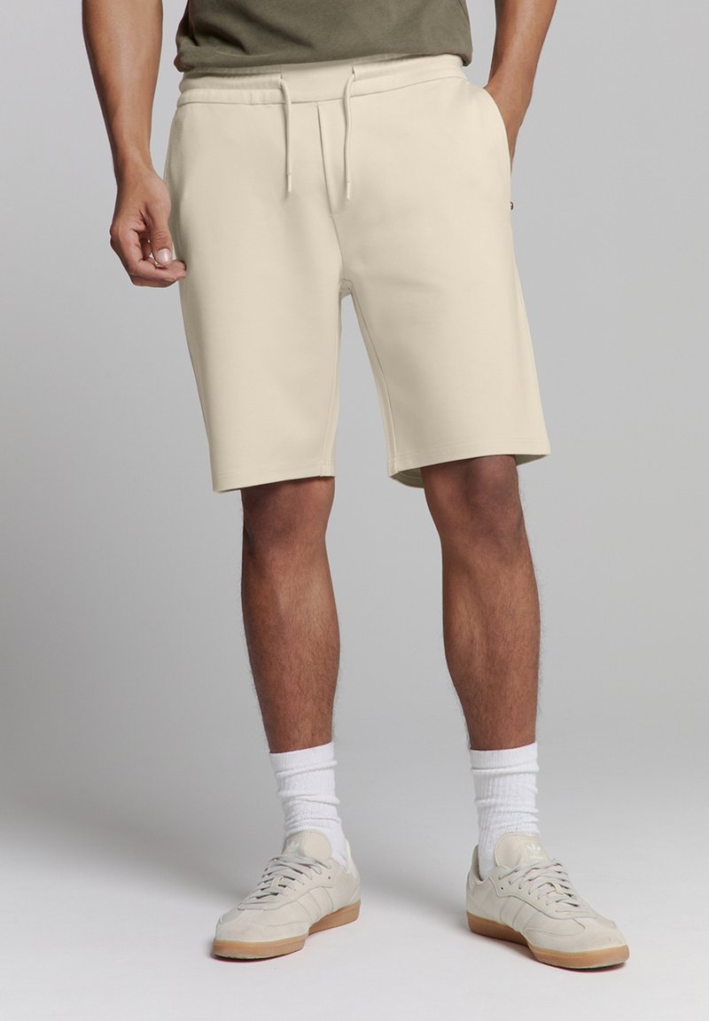 Beige sportshorts gemaakt van zachte stof, met een elastische tailleband en koord, gedragen met witte crew-sokken en lichte sneakers.