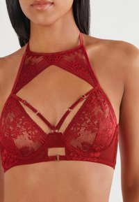 Kvinde iført en rød blonde halterneck bralette med justerbare stropper og blomster mønster mod en simpel lys baggrund.
