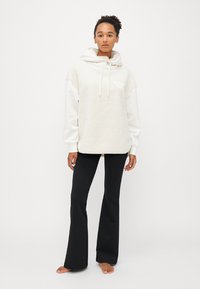 Felpa con cappuccio di pile crema con maniche bianche, lacci, tasca a marsupio e logo discreto. Abbinata a pantaloni neri a campana. Sfondo neutro.