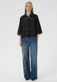 Sort skjorte med knapper og trekvartærmer, der har udskæringsdetaljer og en bølget kant. Kombineret med løse, højtaljede blå jeans.