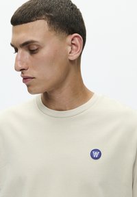 Beige bomulds t-shirt med rund halsudskæring og kortærmer. Har et cirkulært blåt broderet mærke med et hvidt bogstav "W."