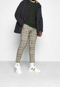 Homme portant une veste noire, un pull vert foncé, un pantalon à carreaux, des chaussettes blanches et des baskets blanches Adidas avec des rayures vertes et jaunes.