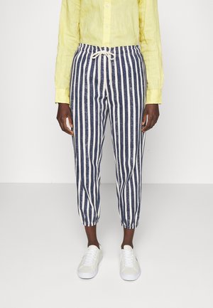 Personne portant un pantalon à rayures verticales bleu marine et blanc avec cordon, une chemise jaune à manches longues et des baskets blanches, debout devant un fond uni.