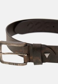 Smeđi kožni pojas s istrošenom teksturom, s srebrnom metalnom kopčom i trokutastim naglaskom. Ime branda "GUESS" ugravirano je na kopči.