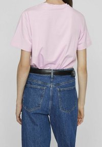 En person står och har på sig en ljusrosa T-shirt och högmidjade blå jeans, sedd bakifrån, med ett svart bälte och en neutral bakgrund.