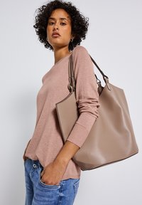 Tote bag aus weichem, strukturiertem beigefarbenem Leder mit doppelten Griffen. Tragend zu einem schlichten rosa Pullover, der ein minimalistisches Design präsentiert.