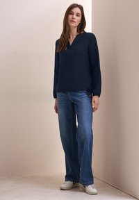 Nagelnieuwe marineblauwe lange mouw top met een V-hals, van textuurstof, gecombineerd met wijdvallende blauwe jeans en witte sneakers met rode accenten.