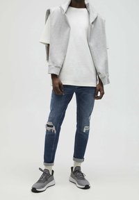 Grey sleeveless hoodie over a white t-shirt, paired with ripped dark blue jeans and grey sneakers. Smooth textures and relaxed fit.