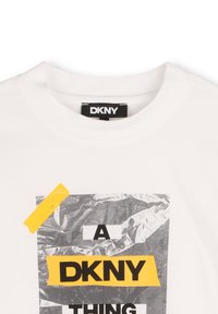 T-shirt blanc en coton avec un design graphique avec du foil argenté et un texte en noir et jaune audacieux, encolure ronde et étiquette DKNY.
