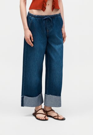 Femeie purtând blugi din denim albastru închis cu talie largă și tiv, top scurt portocaliu și sandale maro cu barete, stând pe fundal alb.