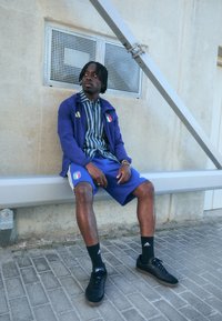 Mėlynas sportinis švarkas su Italijos vėliava, juostuotas marškinėliai, mėlynos šortai, juodos kojinės ir sportbačiai. Sėdi ant suolelio prie tekstūruotų sienų.