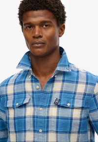 Camicia a scacchi blu e gialla con due tasche sul petto, colletto con bottoni e patch del logo sulla tasca sinistra. Tessuto testurizzato.
