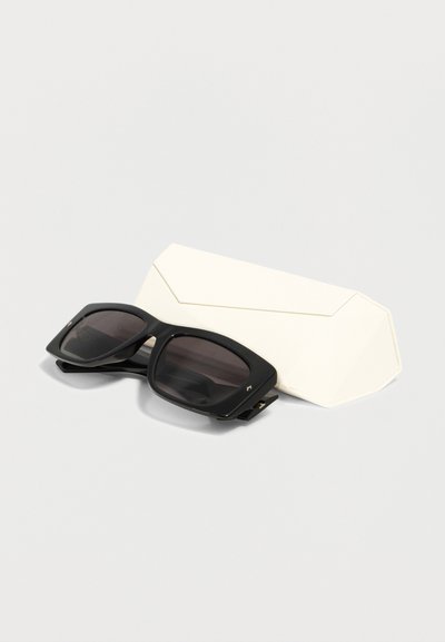 Μαύρα ορθογώνια γυαλιά ηλίου με σκούρους φακούς που ακουμπούν μπροστά από μια κλειστή λευκή γεωμετρική σκληρή θήκη σε ανοιχτό φόντο.
