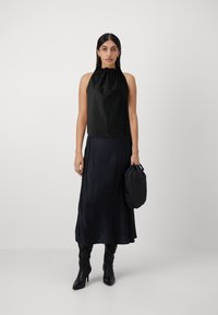 Top nero a halter con scollo arricciato abbinato a una lunga gonna nera fluente. La modella tiene in mano una piccola borsa nera, indossando stivali neri fino al ginocchio.