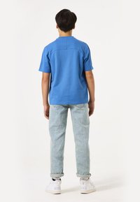 Camisa azul de manga corta con corte recto, combinada con jeans de mezclilla azul claros y zapatillas blancas con un logo visible.