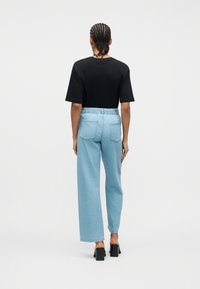 Άτομο με πλεξούδες, φοράει μαύρο μπλουζάκι, ανοιχτόχρωμα τζιν ψηλόμεσα και μαύρα παπούτσια με τακούνι, στέκεται με την πλάτη γυρισμένη σε λευκό φόντο.