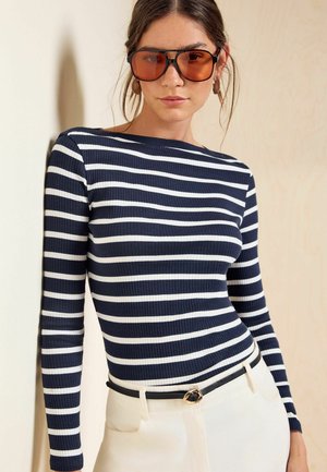Woman wearing a navy and white striped long-sleeve top, white pants with a black belt, and large brown-tinted sunglasses.