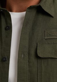 Camisa de lino verde oliva con cuello button-down, botones negros y un bolsillo en el pecho que presenta un detalle de ala bordado. Por dentro se muestra una camisa blanca debajo.