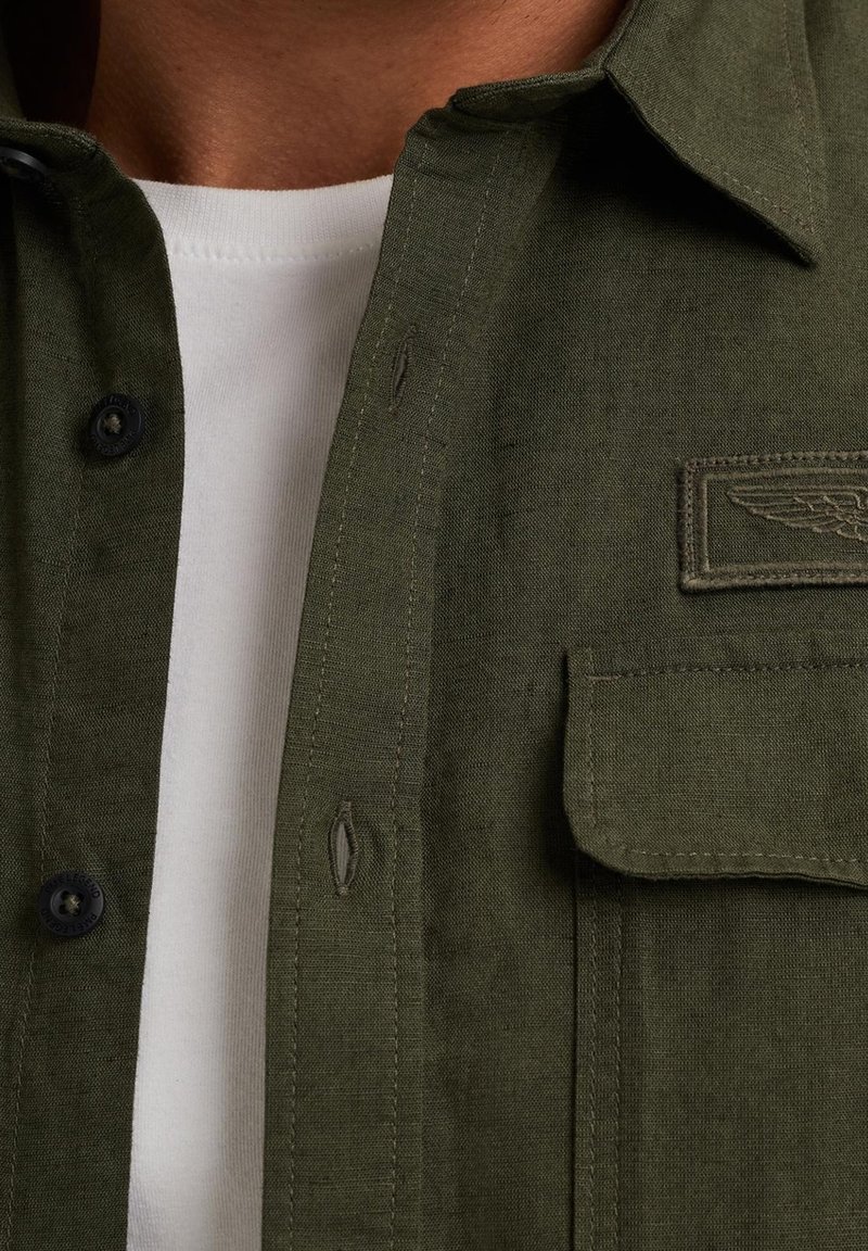 Camisa de lino verde oliva con cuello button-down, botones negros y un bolsillo en el pecho que presenta un detalle de ala bordado. Por dentro se muestra una camisa blanca debajo.