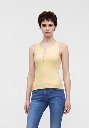 Kvinde med kort krøllet hår iført en gul knapbluset ærmeløs top og blå jeans står mod en ensfarvet lys baggrund.