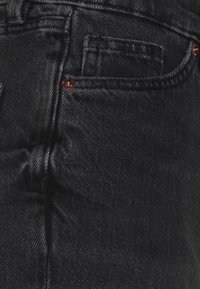 Gros plan d'une poche de jean en denim noir avec des coutures visibles et des rivets en cuivre sur les bords de la poche.