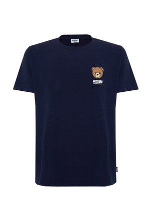 Man met tatoeages die een donkerblauw teddybeer-logo t-shirt draagt, donkere spijkerbroek en bruine bootschoenen, staand tegen een egale lichte achtergrond.