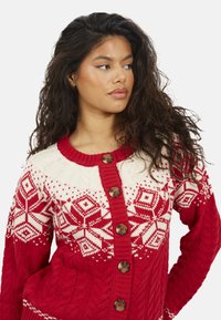 Cardigan lavorato a maglia rosso e crema con motivo a fiocco di neve, caratterizzato da una scollatura rotonda, trama a trecce e cinque grandi bottoni marroni.