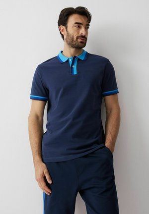 Mężczyzna stoi oparty o ścianę, ubrany w granatową koszulkę polo z jasnoniebieskim kołnierzykiem i akcentami na rękawach oraz dopasowane granatowe spodnie.