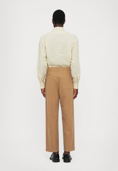 Lys beige lin skjorte med lange ærmer, parret med brune vide bukser. Svarte sko fuldfører outfittet. Synlige baglommer på bukserne.
