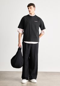 T-shirt preta oversize com mangas curtas, detalhe do logótipo; combinada com calças largas pretas e sapatilhas brancas. Segura uma bolsa de tecido preta.