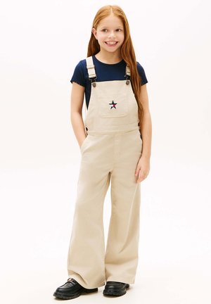 Naeratav tüdruk pikkade punaste juustega, kandes beežid töösärke tähepaelaga, tumesinist lühikese varrukaga särki ja musti kingi, seisab käed taskutes.