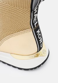 Złote i beżowe dzianinowe sneakersy z gładkimi akcentami, czarno-białymi detalami z logo oraz błyszczącą złotą komponentą na pięcie. Guma podeszwy zapewniająca przyczepność.