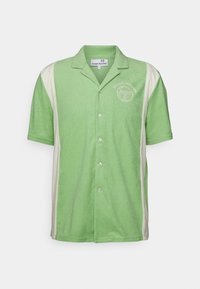 Camisa verde de manga corta con botones y cuello, que presenta acentos crema a los lados y un logo en el pecho izquierdo. Textura suave.