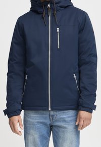 Giacca impermeabile blu navy con zip frontale, cappuccio regolabile e due tasche con zip. Presenta polsini elastici e una texture liscia.