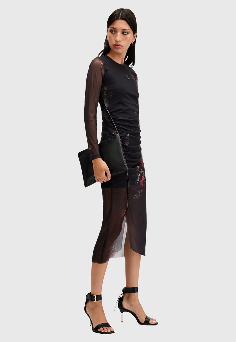 Vestido negro con frunces, mangas largas transparentes y acentos florales; de longitud media con una abertura lateral. Combinado con un bolso negro de mano y sandalias de tacón alto.