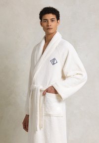 Accappatoio bianco in spugna con colletto a scialle, cintura, e tasca frontale. Presenta un logo ricamato a forma di diamante sul petto.