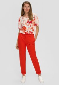 Bloemmotief top in crème, rood en blauw; gecombineerd met rode, tapered pants. Model draagt witte sneakers, met zichtbare manchet-details op de broek.