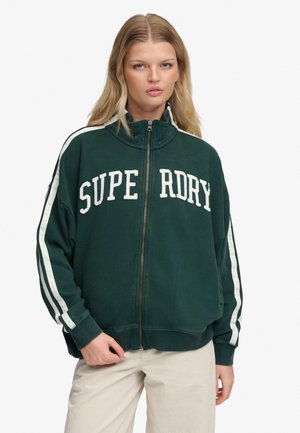 Młoda kobieta o długich blond włosach, ubrana w ciemnozieloną kurtkę zapinaną na zamek, z białymi paskami na rękawach i napisem "SUPERDRY" z przodu.