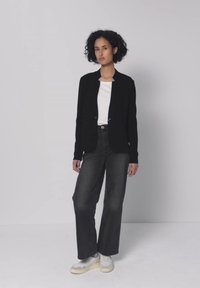 Frau mit lockigem Haar, die eine schwarze Jacke, ein weißes Hemd, weite schwarze Jeans und weiße Sneakers trägt, steht vor einem schlichten hellen Hintergrund.