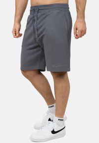 Grijze sweatshorts met drawstring van textuurstof, voorzien van zijzakken en een klein geborduurd logo aan de zoom. Gekoombineerd met witte sneakers.