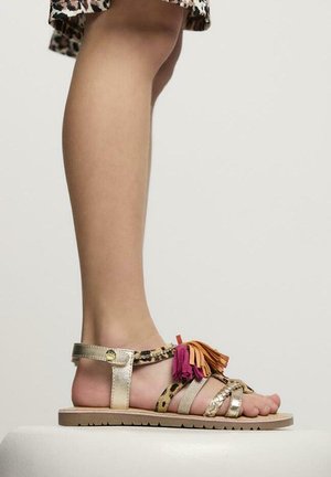 Sandalias metálicas doradas con borlas multicolores, detalles de estampado de leopardo y múltiples tiras, sobre un fondo neutro.