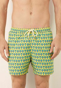 Costume da bagno con uno sfondo giallo brillante e un motivo a cactus ripetuto in blu e verde. Vita con coulisse e dettagli bianchi.