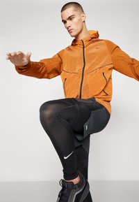 Lichtgewicht oranje zip-up jack met zwarte accenten, gecombineerd met aansluitende zwarte leggings met textuurpanelen en witte logo-details.