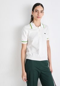 Witte korte mouwen polo met groene en gele accenten op de kraag en mouwen, met drie knopen en een ribbelrand. Lacoste-logo aanwezig.