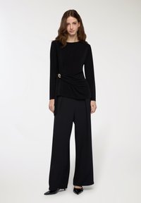 Maglietta nera a maniche lunghe con un design attorcigliato in vita e un accento dorato, abbinata a pantaloni neri a gamba larga e scarpe nere a punta.