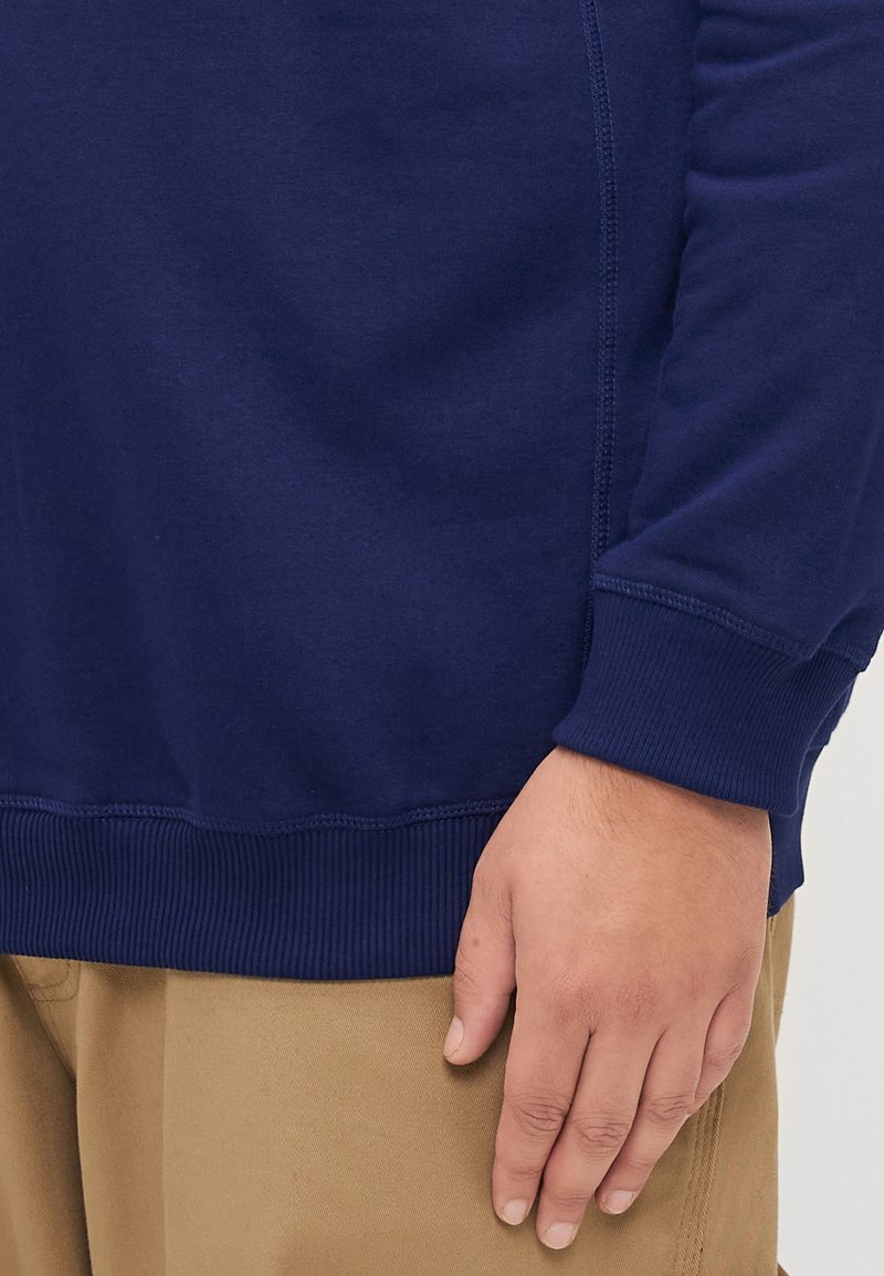 Marineblauwe sweatshirt met ribgebreide boorden en onderzoom, met platte naden en een zachte textuur. De lichtbruine hand van het model rust op beige broek.