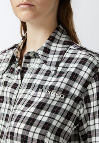 Chemise à carreaux noir et blanc avec des accents roses et gris. Comprend un col, une poche et un logo "PINKO" sur la poitrine.