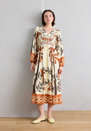 Kvinde iført en langærmet, cremet midi-kjole med orange og brune blomstrede og abstrakte mønstre, iført gule flade sko.