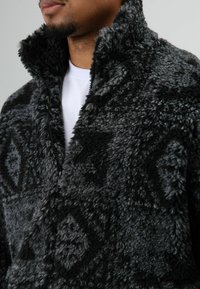 Fleecevest in zwart en grijs met een gestructureerd, pluizig oppervlak, voorzien van een hoge kraag en een ritssluiting aan de voorkant, versierd met ruitpatronen.